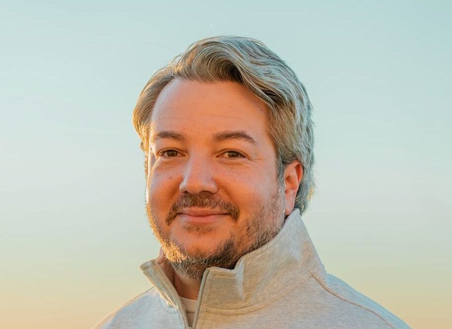 A man with light brown hair and a short beard is wearing a light gray jacket, standing outdoors with a clear sky in the background.