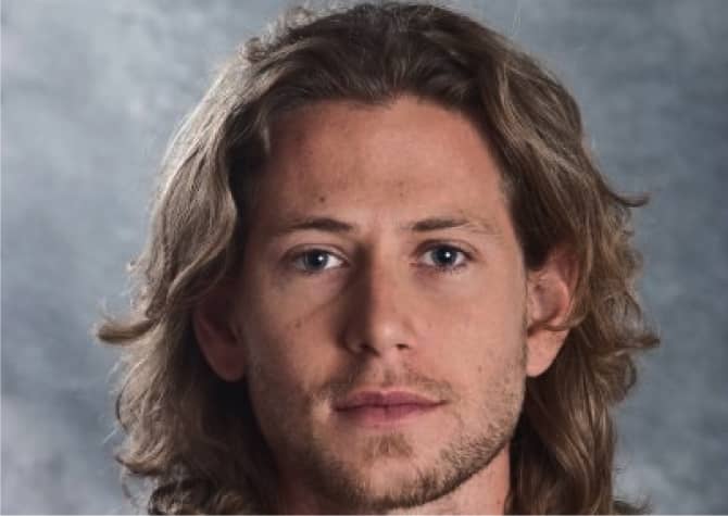 A man with long wavy brown hair, light facial hair, and blue eyes looks directly at the camera against a gray, textured background.
